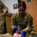A U.S. Air Force medical personnel member wearing OCP (Operational Camouflage Pattern) uniform and blue nitrile gloves is performing a medical training exercise on a simulated casualty lying on the floor. The provider, whose name tape reads 'GONZALEZ,' appears to be conducting an airway assessment or nasopharyngeal airway (NPA) insertion procedure on a person lying supine. A second uniformed individual is visible in the background, suggesting a group training scenario.