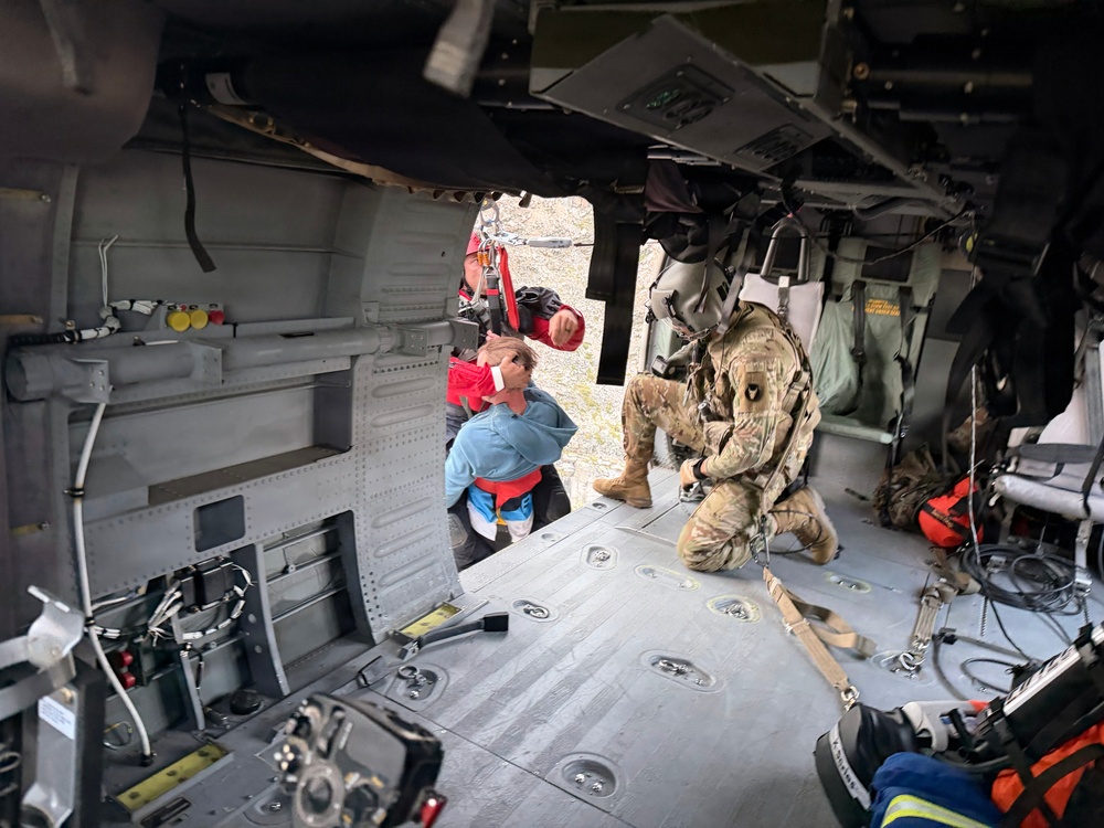 The image shows the interior of a military helicopter during what appears to be a search and rescue operation. A soldier in multicam/OCP uniform and flight helmet is kneeling inside the aircraft near the open door, assisting a civilian being brought aboard. A rescue worker in a red vest is visible at the door opening, helping load the individual.