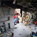 The image shows the interior of a military helicopter during what appears to be a search and rescue operation. A soldier in multicam/OCP uniform and flight helmet is kneeling inside the aircraft near the open door, assisting a civilian being brought aboard. A rescue worker in a red vest is visible at the door opening, helping load the individual.
