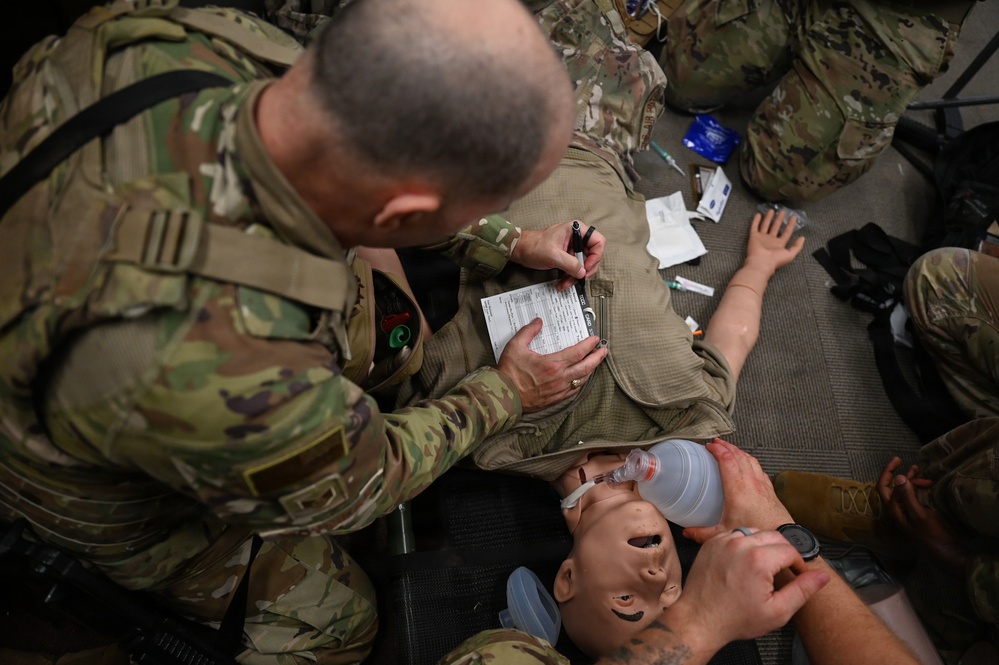 Military personnel in MultiCam/OCP camouflage uniforms are conducting a tactical combat casualty care (TCCC) training exercise. A soldier is filling out what appears to be a casualty card/TCCC card on a medical training mannequin that has a bag-valve mask (BVM) applied to its face. Various medical supplies are visible scattered around the training area.