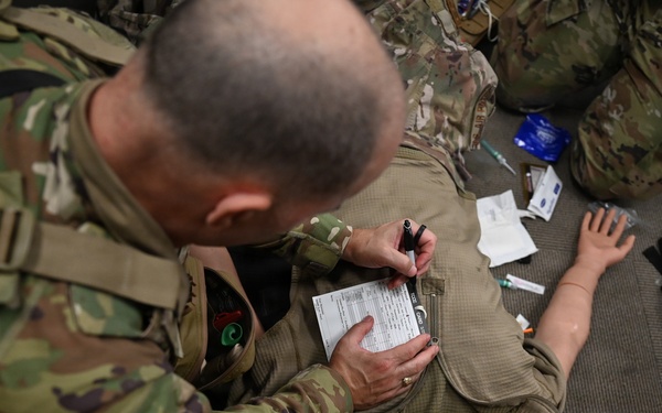 Military personnel in MultiCam/OCP camouflage uniforms are conducting a tactical combat casualty care (TCCC) training exercise. A soldier is filling out what appears to be a casualty card/TCCC card on a medical training mannequin that has a bag-valve mask (BVM) applied to its face. Various medical supplies are visible scattered around the training area.