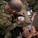 Military personnel in MultiCam/OCP camouflage uniforms are conducting a tactical combat casualty care (TCCC) training exercise. A soldier is filling out what appears to be a casualty card/TCCC card on a medical training mannequin that has a bag-valve mask (BVM) applied to its face. Various medical supplies are visible scattered around the training area.