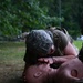 A soldier in US Army ACU (Army Combat Uniform) pattern camouflage is performing rescue breathing/CPR on a medical training mannequin outdoors. The training exercise is taking place on grass with a wooded background. A rifle is visible on the ground nearby, indicating this is a tactical combat casualty care (TCCC) training scenario.