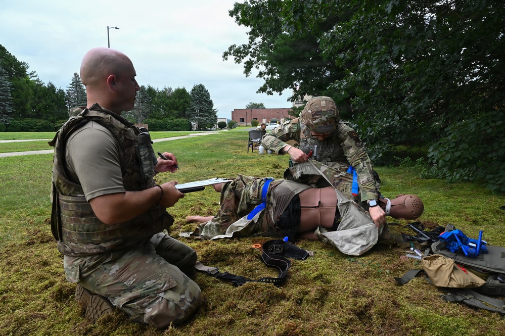 Military personnel are conducting a tactical combat casualty care (TCCC) training exercise outdoors on a grassy area. One soldier is treating a medical training mannequin dressed in military uniform while another soldier kneels nearby holding a clipboard, apparently evaluating or scoring the performance. Various medical training supplies and equipment are scattered on the ground around them.