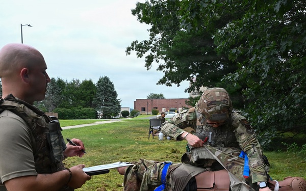 Military personnel are conducting a tactical combat casualty care (TCCC) training exercise outdoors on a grassy area. One soldier is treating a medical training mannequin dressed in military uniform while another soldier kneels nearby holding a clipboard, apparently evaluating or scoring the performance. Various medical training supplies and equipment are scattered on the ground around them.