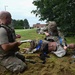 Military personnel are conducting a tactical combat casualty care (TCCC) training exercise outdoors on a grassy area. One soldier is treating a medical training mannequin dressed in military uniform while another soldier kneels nearby holding a clipboard, apparently evaluating or scoring the performance. Various medical training supplies and equipment are scattered on the ground around them.