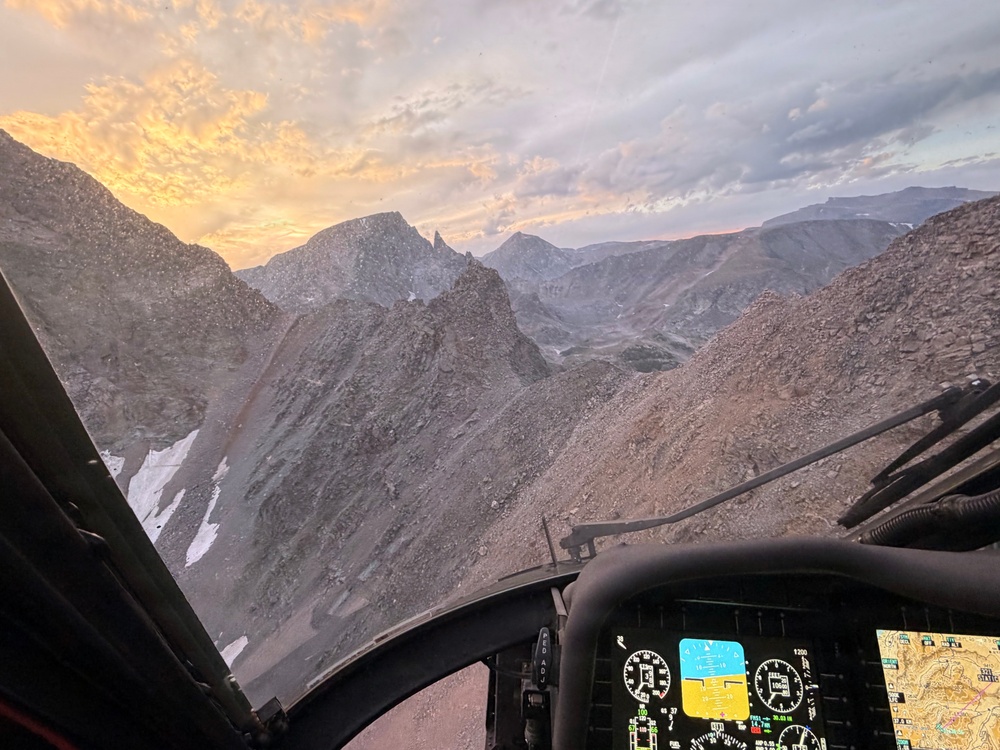 A photograph taken from inside a helicopter cockpit flying through a rugged, high-altitude mountain range at sunset or sunrise. The instrument panel and navigation displays are visible in the foreground, while dramatic rocky peaks, scree slopes, and patches of snow fill the view through the windshield. The warm orange and golden sky contrasts with the grey, barren terrain below.