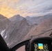 A photograph taken from inside a helicopter cockpit flying through a rugged, high-altitude mountain range at sunset or sunrise. The instrument panel and navigation displays are visible in the foreground, while dramatic rocky peaks, scree slopes, and patches of snow fill the view through the windshield. The warm orange and golden sky contrasts with the grey, barren terrain below.