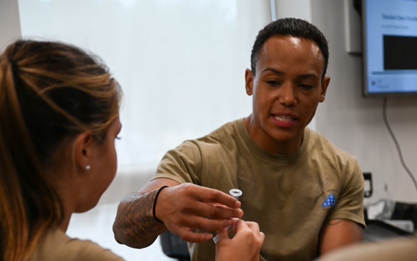 Two military personnel in olive/tan uniforms are engaged in what appears to be a medical training or treatment scenario. One individual is seated on an examination table while the other handles a small medical vial or connector, suggesting IV or injection training. A monitor is visible in the background, consistent with a medical training facility.