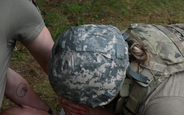 A soldier wearing ACU pattern helmet and OCP/Multicam plate carrier is performing tactical combat casualty care (TCCC) on a simulated casualty during a field training exercise. The provider appears to be performing a needle decompression or chest seal application on the patient's chest. Medical training materials and simulated blood are visible, indicating this is a realistic medical training scenario.