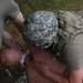A soldier wearing ACU pattern helmet and OCP/Multicam plate carrier is performing tactical combat casualty care (TCCC) on a simulated casualty during a field training exercise. The provider appears to be performing a needle decompression or chest seal application on the patient's chest. Medical training materials and simulated blood are visible, indicating this is a realistic medical training scenario.