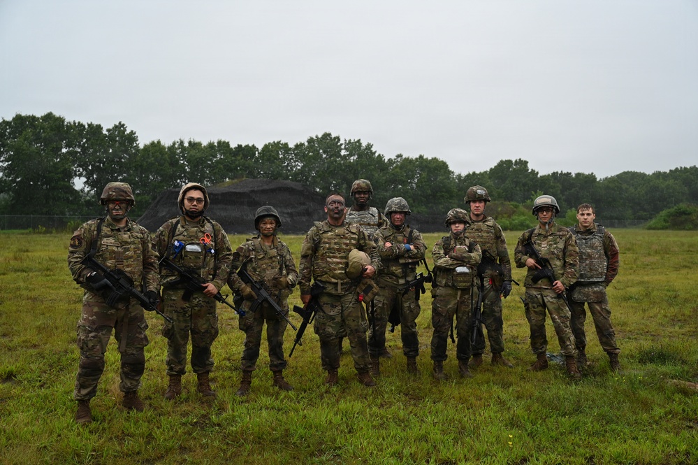 A group of approximately nine military personnel in full combat gear pose for a group photograph on a grassy field. They are wearing camouflage uniforms, tactical vests, and helmets, with several carrying rifles. The setting appears to be a training area, evidenced by a dirt berm structure visible in the background.