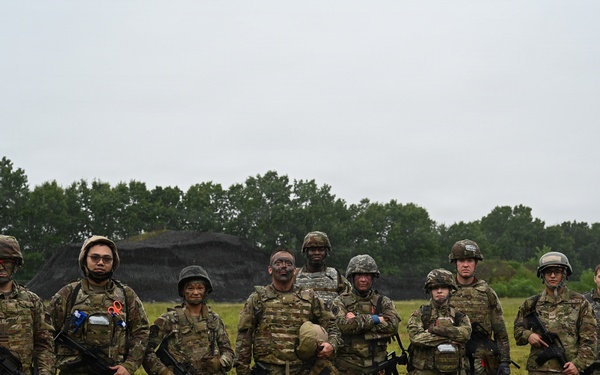 A group of approximately nine military personnel in full combat gear pose for a group photograph on a grassy field. They are wearing camouflage uniforms, tactical vests, and helmets, with several carrying rifles. The setting appears to be a training area, evidenced by a dirt berm structure visible in the background.