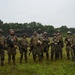 A group of approximately nine military personnel in full combat gear pose for a group photograph on a grassy field. They are wearing camouflage uniforms, tactical vests, and helmets, with several carrying rifles. The setting appears to be a training area, evidenced by a dirt berm structure visible in the background.