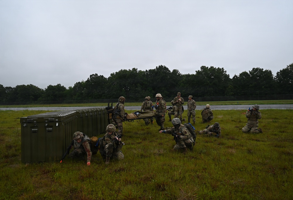 A group of military personnel in camouflage uniforms are conducting a field training exercise on a grassy open area adjacent to what appears to be a runway or tarmac. Several soldiers are in low crouching or prone positions while others are standing and carrying a litter/stretcher, suggesting a casualty evacuation drill. A large green military equipment container is visible on the left side of the scene.