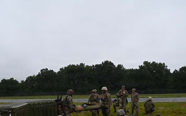 A group of military personnel in camouflage uniforms are conducting a field training exercise on a grassy open area adjacent to what appears to be a runway or tarmac. Several soldiers are in low crouching or prone positions while others are standing and carrying a litter/stretcher, suggesting a casualty evacuation drill. A large green military equipment container is visible on the left side of the scene.