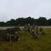 A group of military personnel in camouflage uniforms are conducting a field training exercise on a grassy open area adjacent to what appears to be a runway or tarmac. Several soldiers are in low crouching or prone positions while others are standing and carrying a litter/stretcher, suggesting a casualty evacuation drill. A large green military equipment container is visible on the left side of the scene.