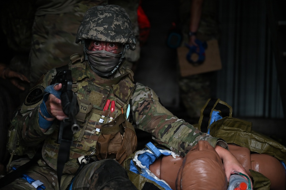 A soldier in full combat gear and camouflage uniform is performing tactical combat casualty care (TCCC) on what appears to be a training mannequin or simulated casualty. The soldier is holding a weapon in one hand while attending to the simulated patient with the other, suggesting a 'care under fire' training scenario. Red simulated blood markings are visible on the soldier's face, and training markers are attached to their vest.