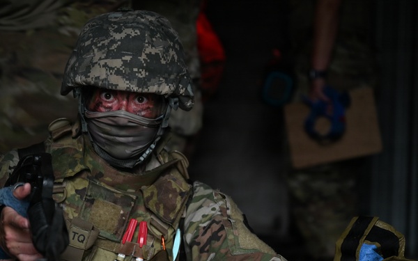 A soldier in full combat gear and camouflage uniform is performing tactical combat casualty care (TCCC) on what appears to be a training mannequin or simulated casualty. The soldier is holding a weapon in one hand while attending to the simulated patient with the other, suggesting a 'care under fire' training scenario. Red simulated blood markings are visible on the soldier's face, and training markers are attached to their vest.