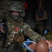A soldier in full combat gear and camouflage uniform is performing tactical combat casualty care (TCCC) on what appears to be a training mannequin or simulated casualty. The soldier is holding a weapon in one hand while attending to the simulated patient with the other, suggesting a 'care under fire' training scenario. Red simulated blood markings are visible on the soldier's face, and training markers are attached to their vest.