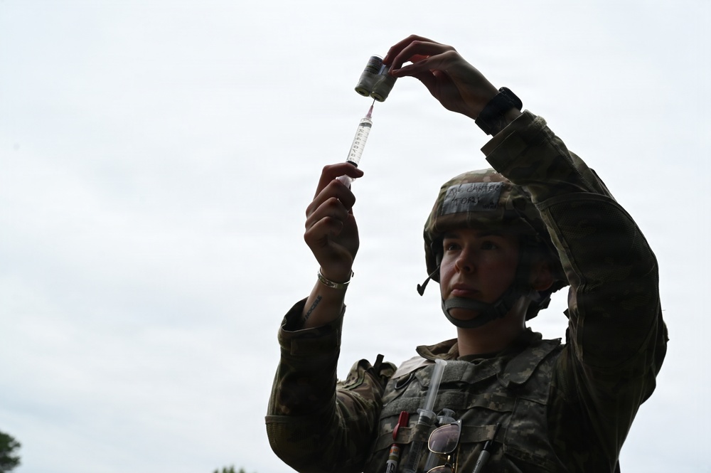 A female soldier in military combat uniform and helmet is drawing medication from a vial into a syringe, holding both items raised above her head to check the dosage. She appears to be a military medic or combat medical specialist performing a standard medical preparation procedure outdoors. The low-angle shot emphasizes her focused concentration on the task.