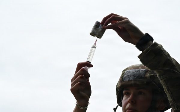 A female soldier in military combat uniform and helmet is drawing medication from a vial into a syringe, holding both items raised above her head to check the dosage. She appears to be a military medic or combat medical specialist performing a standard medical preparation procedure outdoors. The low-angle shot emphasizes her focused concentration on the task.