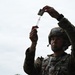 A female soldier in military combat uniform and helmet is drawing medication from a vial into a syringe, holding both items raised above her head to check the dosage. She appears to be a military medic or combat medical specialist performing a standard medical preparation procedure outdoors. The low-angle shot emphasizes her focused concentration on the task.