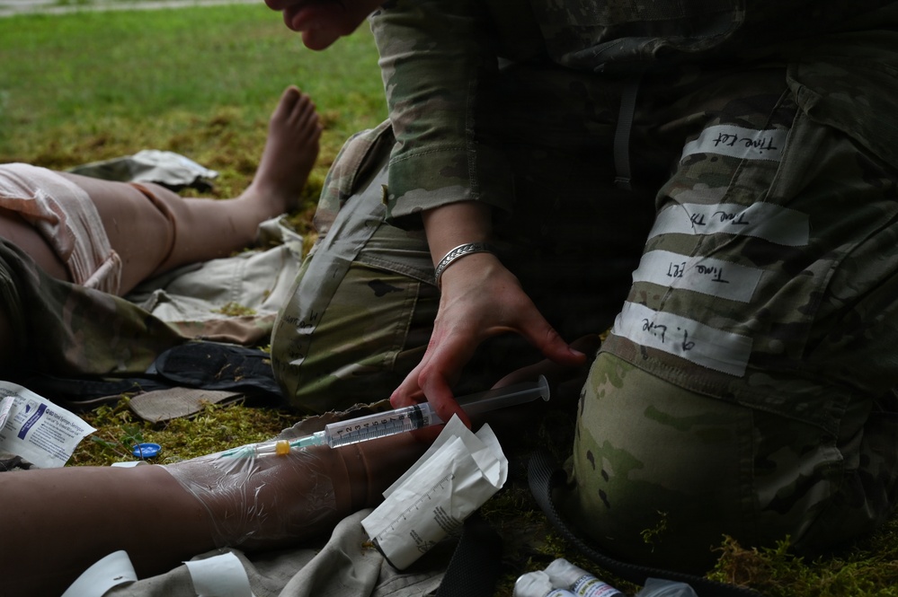 A military medic in OCP (Operational Camouflage Pattern) uniform is administering medical treatment to a casualty on the ground. The medic is handling a syringe near what appears to be a wrapped/bandaged limb, with various medical supplies scattered nearby. White tape with handwritten notations is visible on the medic's leg, consistent with tracking treatment times during a field exercise or real casualty care.