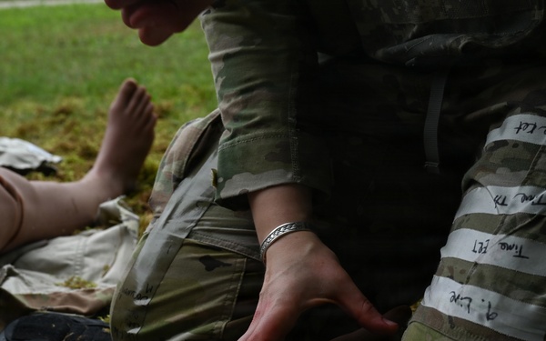 A military medic in OCP (Operational Camouflage Pattern) uniform is administering medical treatment to a casualty on the ground. The medic is handling a syringe near what appears to be a wrapped/bandaged limb, with various medical supplies scattered nearby. White tape with handwritten notations is visible on the medic's leg, consistent with tracking treatment times during a field exercise or real casualty care.