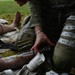 A military medic in OCP (Operational Camouflage Pattern) uniform is administering medical treatment to a casualty on the ground. The medic is handling a syringe near what appears to be a wrapped/bandaged limb, with various medical supplies scattered nearby. White tape with handwritten notations is visible on the medic's leg, consistent with tracking treatment times during a field exercise or real casualty care.