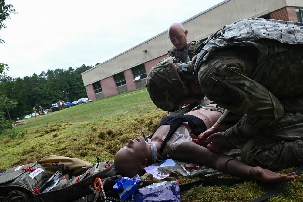 Two military personnel in camouflage uniforms are conducting a combat casualty care training exercise outdoors on a grass field. They are practicing medical procedures on a realistic training mannequin laid on the ground, surrounded by medical supplies and equipment. A brick building and additional personnel are visible in the background.