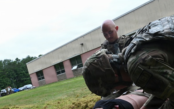Two military personnel in camouflage uniforms are conducting a combat casualty care training exercise outdoors on a grass field. They are practicing medical procedures on a realistic training mannequin laid on the ground, surrounded by medical supplies and equipment. A brick building and additional personnel are visible in the background.