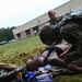 Two military personnel in camouflage uniforms are conducting a combat casualty care training exercise outdoors on a grass field. They are practicing medical procedures on a realistic training mannequin laid on the ground, surrounded by medical supplies and equipment. A brick building and additional personnel are visible in the background.