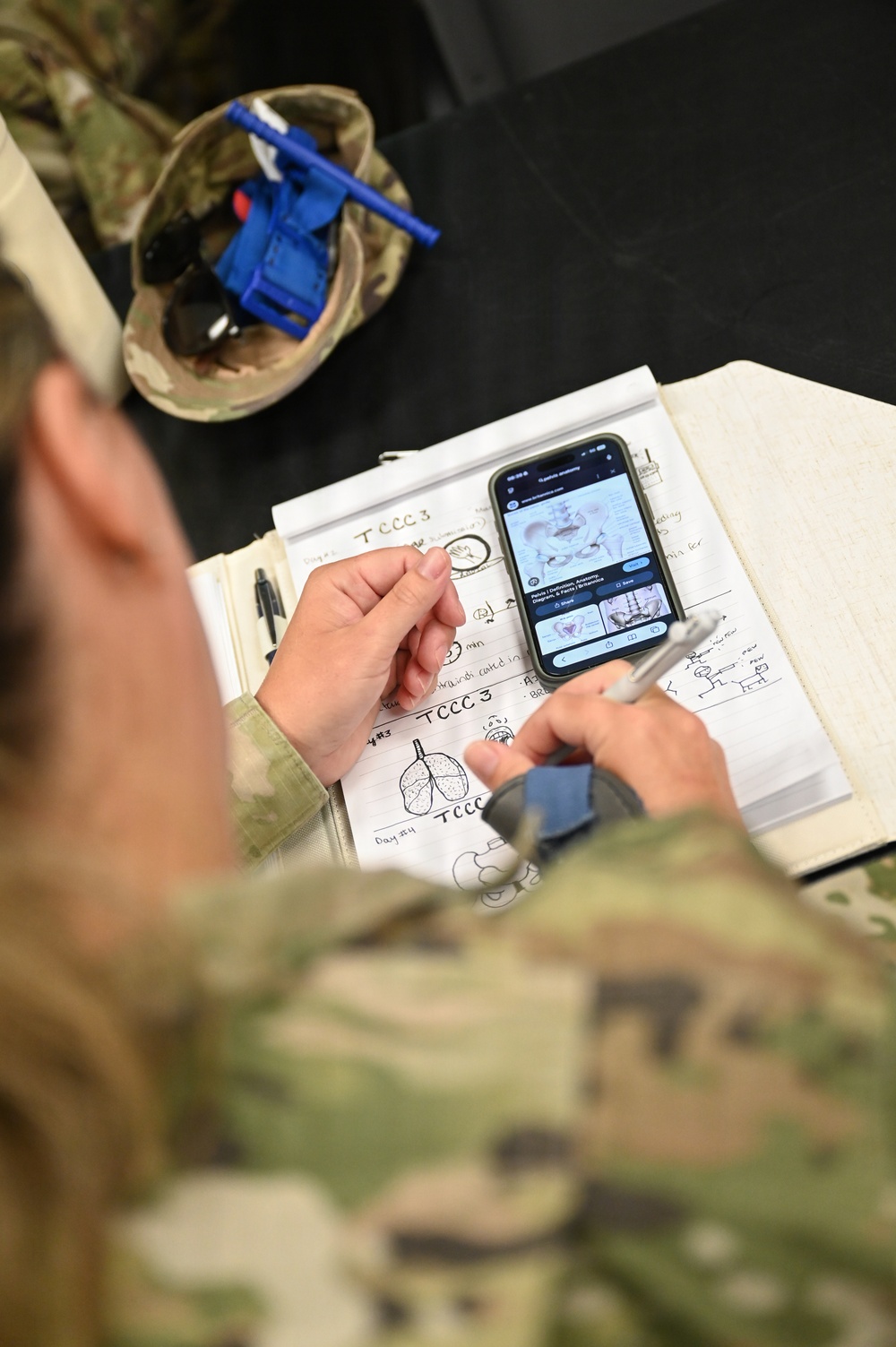 A military service member in camouflage (OCP/MultiCam) uniform is studying medical training materials at a desk. The individual is referencing a smartphone displaying anatomical images while reviewing handwritten notes labeled 'TCCC 3' (Tactical Combat Casualty Care, Day 3). The notes include hand-drawn anatomical diagrams including lungs and other body structures.