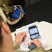 A military service member in camouflage (OCP/MultiCam) uniform is studying medical training materials at a desk. The individual is referencing a smartphone displaying anatomical images while reviewing handwritten notes labeled 'TCCC 3' (Tactical Combat Casualty Care, Day 3). The notes include hand-drawn anatomical diagrams including lungs and other body structures.