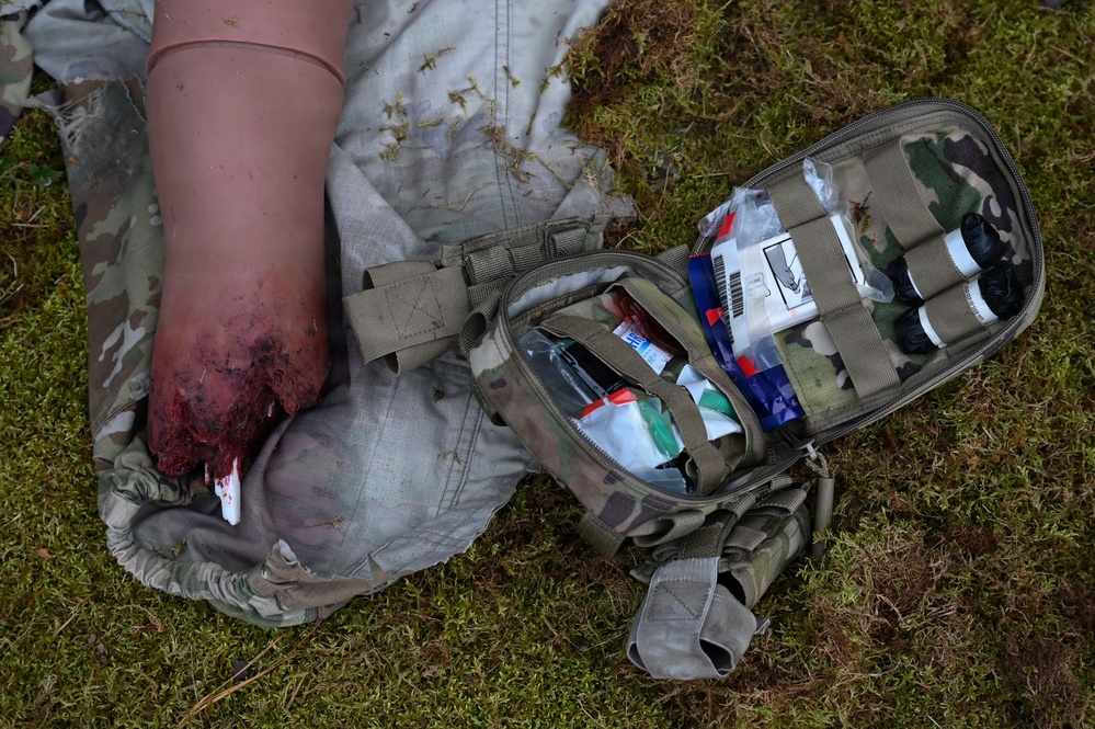 The image depicts a tactical combat casualty care (TCCC) scenario, likely a training exercise, showing a simulated wounded limb with a tourniquet applied and visible simulated blood. An open military-style Individual First Aid Kit (IFAK) in MultiCam/OCP pattern is positioned nearby on grass, with medical supplies visible inside. The scene is consistent with combat medic or military first aid training.