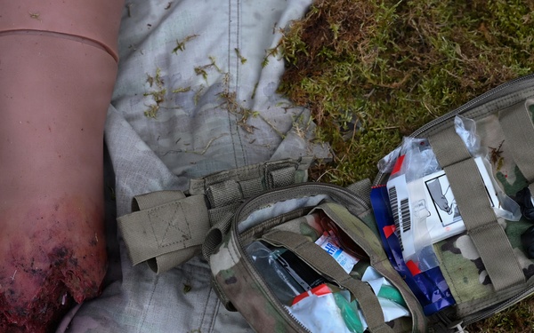 The image depicts a tactical combat casualty care (TCCC) scenario, likely a training exercise, showing a simulated wounded limb with a tourniquet applied and visible simulated blood. An open military-style Individual First Aid Kit (IFAK) in MultiCam/OCP pattern is positioned nearby on grass, with medical supplies visible inside. The scene is consistent with combat medic or military first aid training.