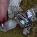 The image depicts a tactical combat casualty care (TCCC) scenario, likely a training exercise, showing a simulated wounded limb with a tourniquet applied and visible simulated blood. An open military-style Individual First Aid Kit (IFAK) in MultiCam/OCP pattern is positioned nearby on grass, with medical supplies visible inside. The scene is consistent with combat medic or military first aid training.