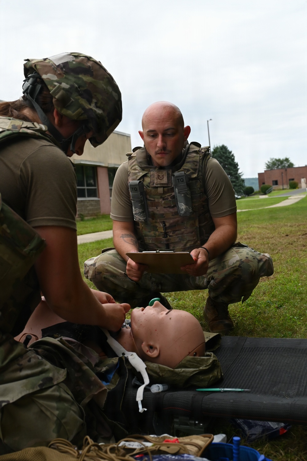 Two military personnel in OCP (Operational Camouflage Pattern) uniforms are conducting a tactical combat casualty care (TCCC) training exercise outdoors on a grassy area. One individual wearing a helmet is performing medical procedures on a training mannequin, while a second individual wearing a tactical vest holds a clipboard, apparently evaluating or scoring the exercise. Medical supplies and equipment are visible on the ground nearby.