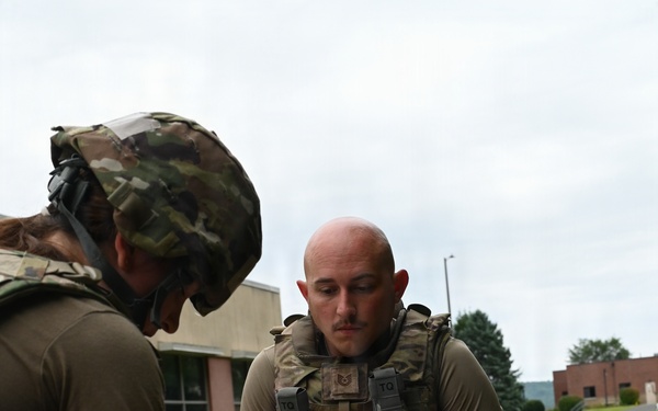 Two military personnel in OCP (Operational Camouflage Pattern) uniforms are conducting a tactical combat casualty care (TCCC) training exercise outdoors on a grassy area. One individual wearing a helmet is performing medical procedures on a training mannequin, while a second individual wearing a tactical vest holds a clipboard, apparently evaluating or scoring the exercise. Medical supplies and equipment are visible on the ground nearby.