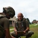 Two military personnel in OCP (Operational Camouflage Pattern) uniforms are conducting a tactical combat casualty care (TCCC) training exercise outdoors on a grassy area. One individual wearing a helmet is performing medical procedures on a training mannequin, while a second individual wearing a tactical vest holds a clipboard, apparently evaluating or scoring the exercise. Medical supplies and equipment are visible on the ground nearby.