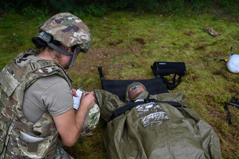 A military medic in full combat gear (helmet, body armor, multicam uniform) is kneeling beside a casualty lying on a litter, taking notes on a small card or form. The casualty is wrapped in an olive-drab emergency blanket/sleeping bag system and appears to be wearing eye protection. The scene appears to be a field medical training exercise conducted on grassy terrain.
