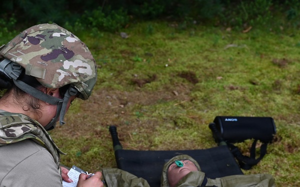 A military medic in full combat gear (helmet, body armor, multicam uniform) is kneeling beside a casualty lying on a litter, taking notes on a small card or form. The casualty is wrapped in an olive-drab emergency blanket/sleeping bag system and appears to be wearing eye protection. The scene appears to be a field medical training exercise conducted on grassy terrain.