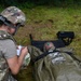 A military medic in full combat gear (helmet, body armor, multicam uniform) is kneeling beside a casualty lying on a litter, taking notes on a small card or form. The casualty is wrapped in an olive-drab emergency blanket/sleeping bag system and appears to be wearing eye protection. The scene appears to be a field medical training exercise conducted on grassy terrain.