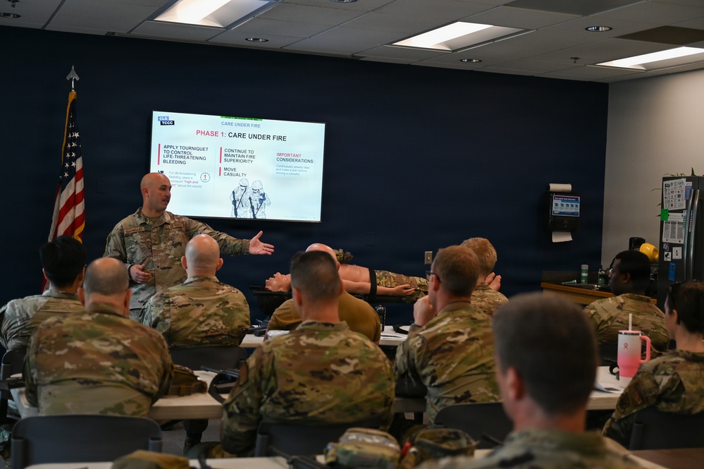 A military instructor is conducting a TCCC (Tactical Combat Casualty Care) training session in a classroom setting. The presentation slide visible on the screen displays 'Phase 1: Care Under Fire,' covering topics such as tourniquet application, maintaining fire superiority, and moving casualties. Multiple uniformed military personnel in camouflage uniforms are seated as students, with one appearing to demonstrate a technique on another.