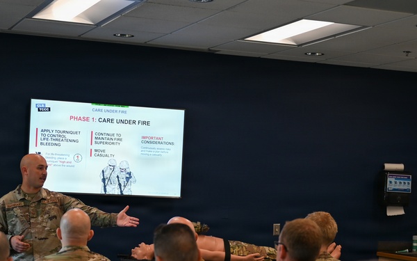 A military instructor is conducting a TCCC (Tactical Combat Casualty Care) training session in a classroom setting. The presentation slide visible on the screen displays 'Phase 1: Care Under Fire,' covering topics such as tourniquet application, maintaining fire superiority, and moving casualties. Multiple uniformed military personnel in camouflage uniforms are seated as students, with one appearing to demonstrate a technique on another.