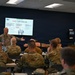 A military instructor is conducting a TCCC (Tactical Combat Casualty Care) training session in a classroom setting. The presentation slide visible on the screen displays 'Phase 1: Care Under Fire,' covering topics such as tourniquet application, maintaining fire superiority, and moving casualties. Multiple uniformed military personnel in camouflage uniforms are seated as students, with one appearing to demonstrate a technique on another.