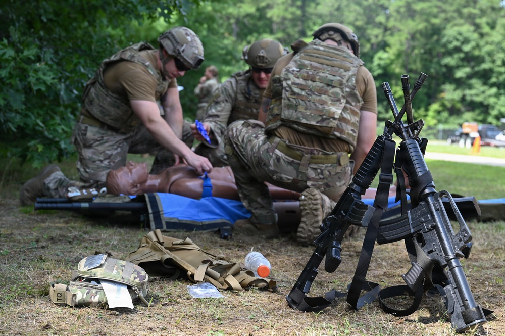 Military personnel in full combat gear and helmets are conducting tactical combat casualty care (TCCC) training on a medical mannequin placed on a blue tarp. Multiple soldiers are practicing trauma treatment procedures while their rifles are stacked together in the foreground. Medical supplies, tactical vests, and equipment are scattered on the ground nearby.