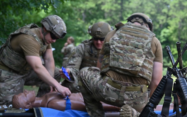 Military personnel in full combat gear and helmets are conducting tactical combat casualty care (TCCC) training on a medical mannequin placed on a blue tarp. Multiple soldiers are practicing trauma treatment procedures while their rifles are stacked together in the foreground. Medical supplies, tactical vests, and equipment are scattered on the ground nearby.