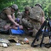 Military personnel in full combat gear and helmets are conducting tactical combat casualty care (TCCC) training on a medical mannequin placed on a blue tarp. Multiple soldiers are practicing trauma treatment procedures while their rifles are stacked together in the foreground. Medical supplies, tactical vests, and equipment are scattered on the ground nearby.